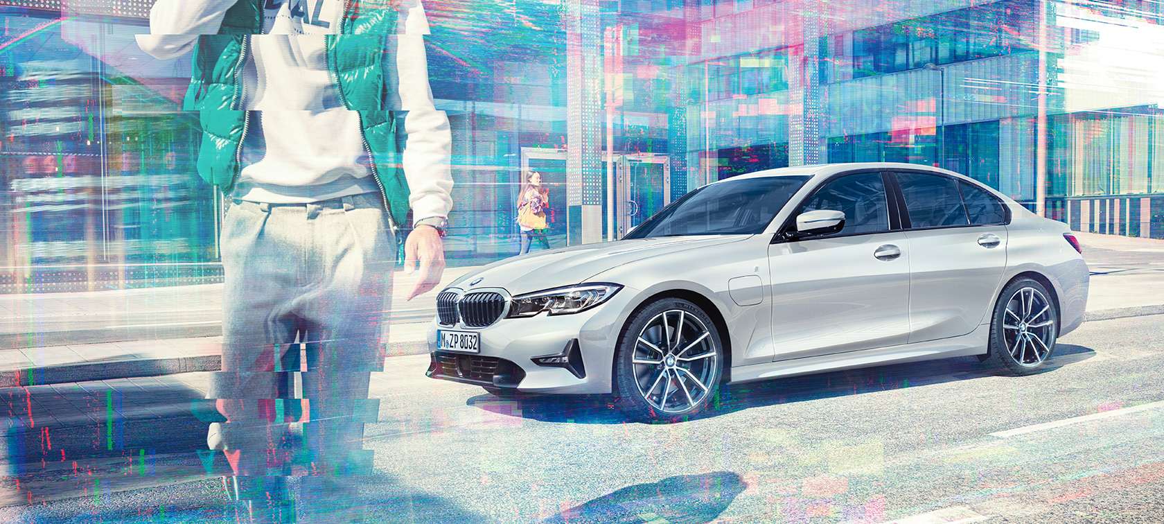 BMW 3-serie Sedan, BMW 330e set fra siden. Stående BMW 330e Sedan set fra siden let drejet, foran glasbygning i urbant miljø med personer i bevægelse.