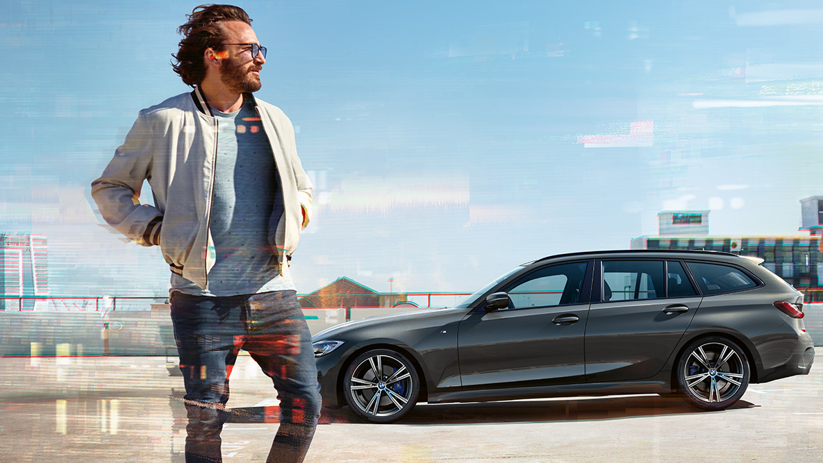 BMW 3-serie Touring (G21) set forfra let drejet, design BMW 3-serie Touring (G21) dravitgrå set forfra let drejet kørende på en bro foran landlige omgivelser