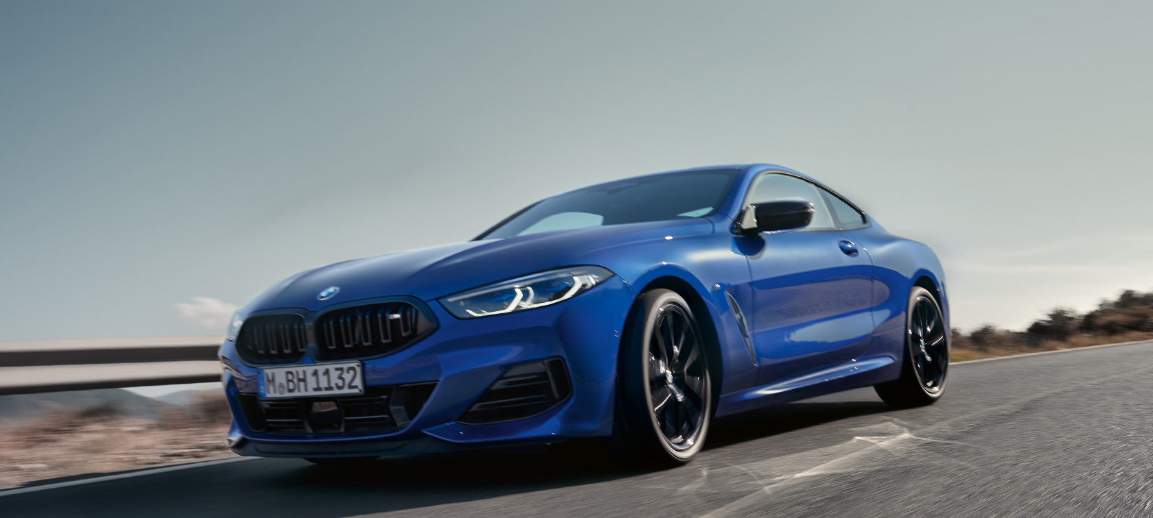 BMW 8-serie Coupé G15 LCI Facelift 2022 BMW M850i xDrive M Portimao Blue metallic set forfra i frøperspektiv kørende BMW 8-serie Coupé G15 LCI Facelift 2022 BMW M850i xDrive M Portimao Blue metallic set forfra i frøperspektiv kørende på landevej foran bakket landskab