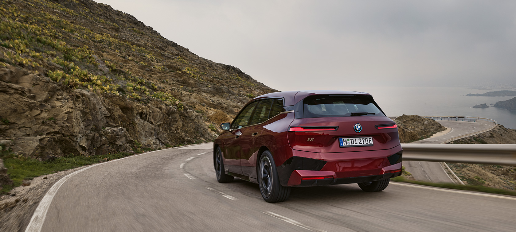 BMW iX i20 2021 elektrisk SUV BMW iX xDrive50 kørende på kystvej BMW iX i20 2021 elektrisk SUV BMW iX xDrive50 Aventurin Red set bagfra kørende på en kystvej