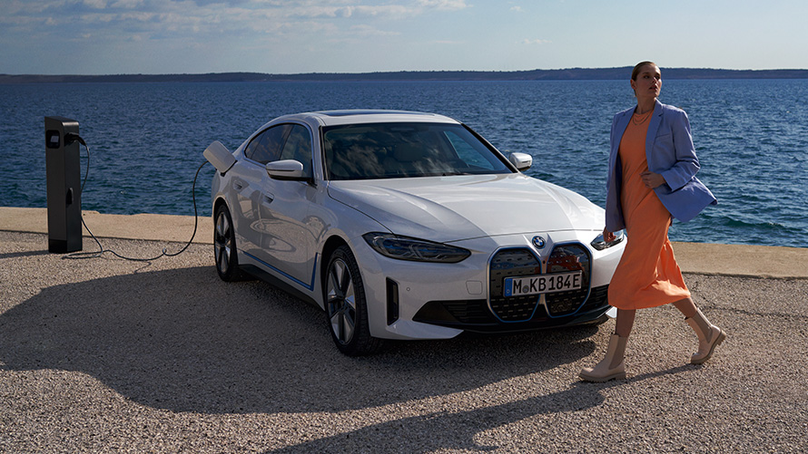 BMW i4 eDrive40 G26 2021 set forfra let drejet ved ladestation Kvinde foran en BMW i4 eDrive40 G26 2021 mineralhvid set forfra let drejet ved ladestander i nærheden af havet