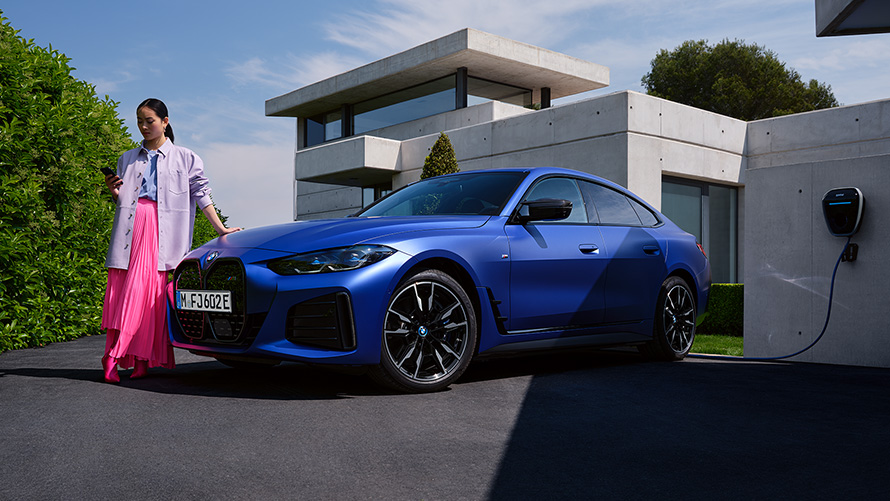 BMW i4 M50 G26 2021 BMW Individual Frozen Portimao Blue metallic set forfra let drejet stående ved siden af en kvinde foran et hus