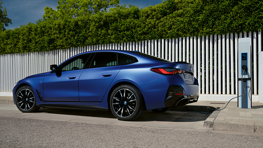 Visning fra siden BMW i4 M50 G26 2021 ved en offentlig hurtigladestation BMW i4 M50 G26 2021 BMW Individual Frozen Portimao Blue metallic set fra siden ved en offentlig hurtigladestation