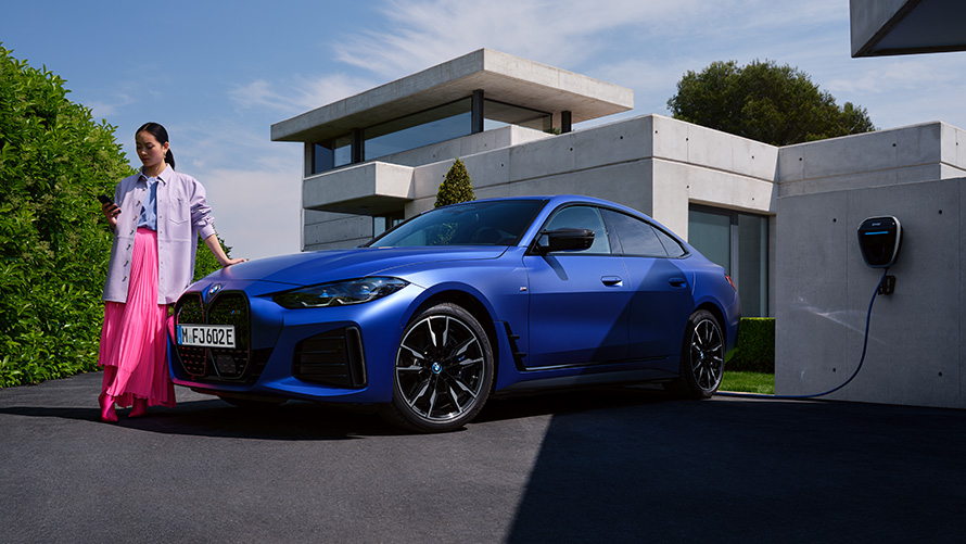 Visning fra siden let drejet BMW i4 M50 G26 2021 med en kvinde foran et hus BMW i4 M50 G26 2021 BMW Individual Frozen Portimao Blue metallic set fra siden let drejet med en kvinde foran et hus