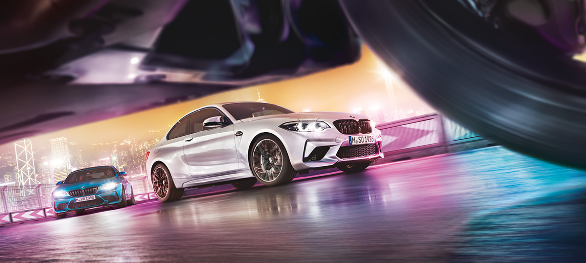 BMW M2 Competition på blank asfalt To BMW M2 Competition på racerbanen set i gadehøjde med storbyens lys som backdrop.