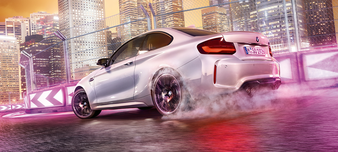 BMW M2 Competition under acceleration Hvid BMW M2 Competition, der drifter på racerbanen med storbyens lys som backdrop.
