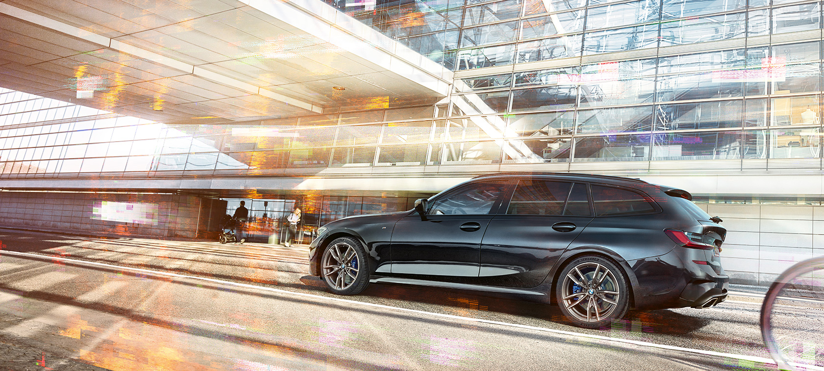 BMW M340i xDrive Touring, billede taget fra siden under kørsel BMW M340i xDrive Touring, billede taget fra siden under kørsel foran en urban glasbygning