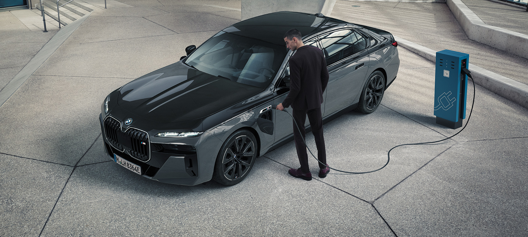 BMW M760e xDrive Sedan G70 M Performance pakke BMW Individual two tone-lakering Sapphire Black metallic BMW Individual Dravit Grey metallic set forfra let drejet mand med ladekabel ved siden af ladestation