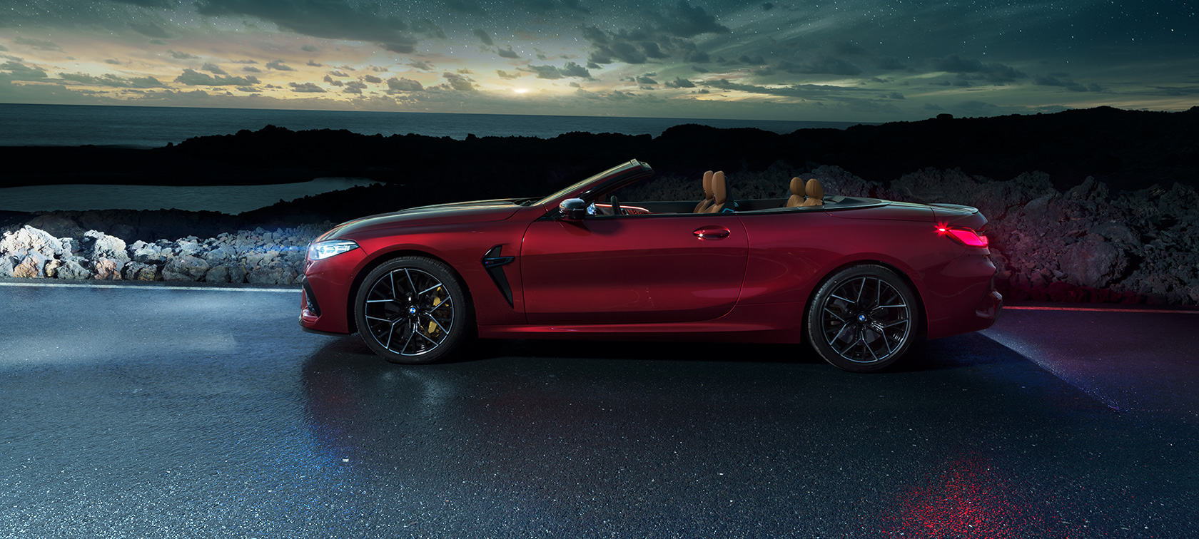 BMW M8 Competition Cabriolet set fra siden BMW M8 Competition Cabriolet i Motegi Red metallic med åben kaleche set fra siden, parkeret på en kystvej under en stjernehimmel.