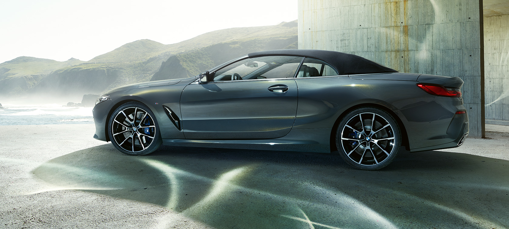 BMW M850i xDrive Cabriolet set fra siden. BMW M850i xDrive Cabriolet i Dravit Grey metallic med lukket kaleche, set fra siden foran et højdedrag.