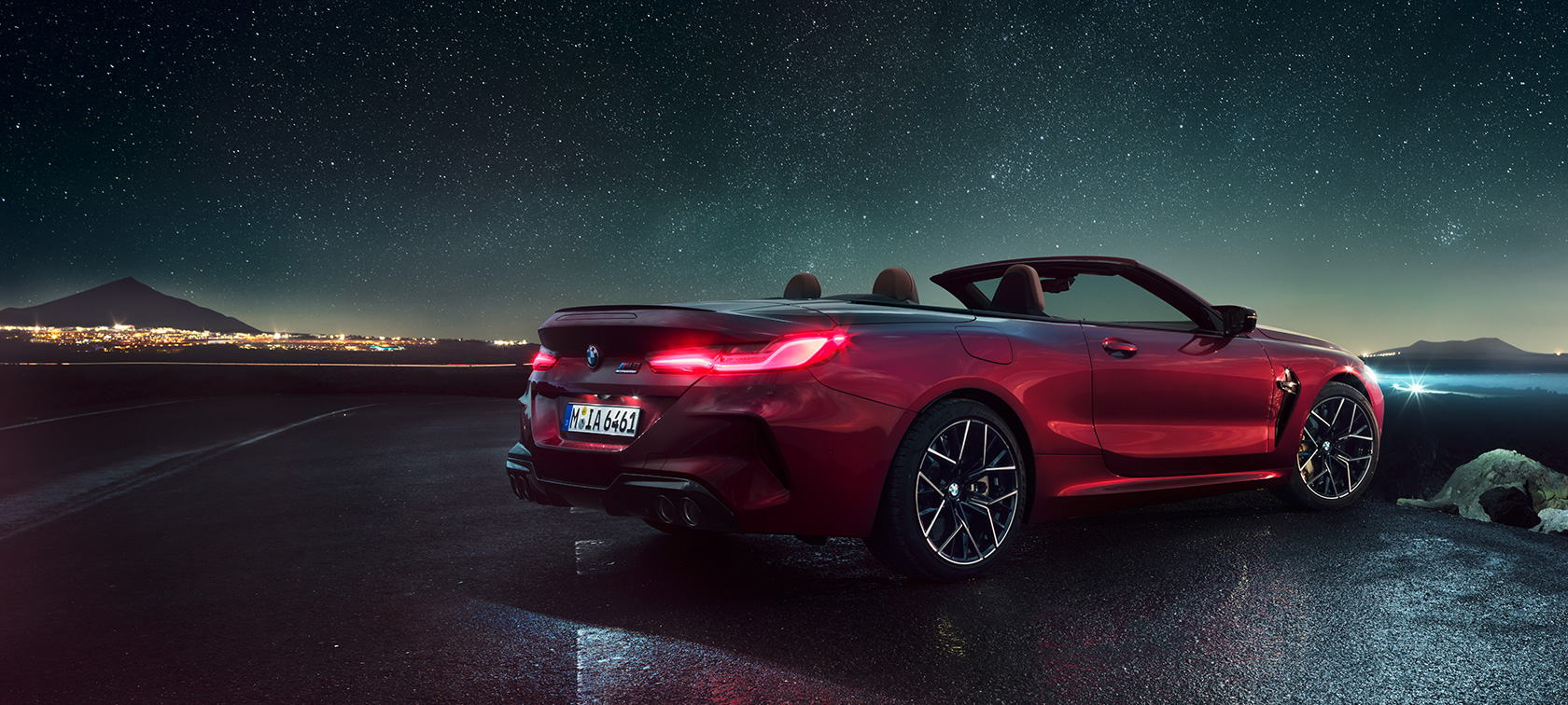 BMW M8 Competition Cabriolet set bagfra let drejet. BMW M8 Competition Cabriolet i Motegi Red metallic med åben kaleche set bagfra let drejet under en stjernehimmel.