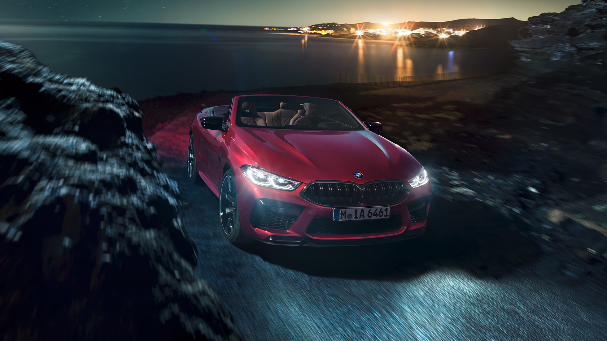 BMW M8 Competition Cabriolet set forfra let drejet. BMW M8 Competition Cabriolet i Motegi Red metallic med åben kaleche set forfra let drejet, kørende om natten på en kystvej.