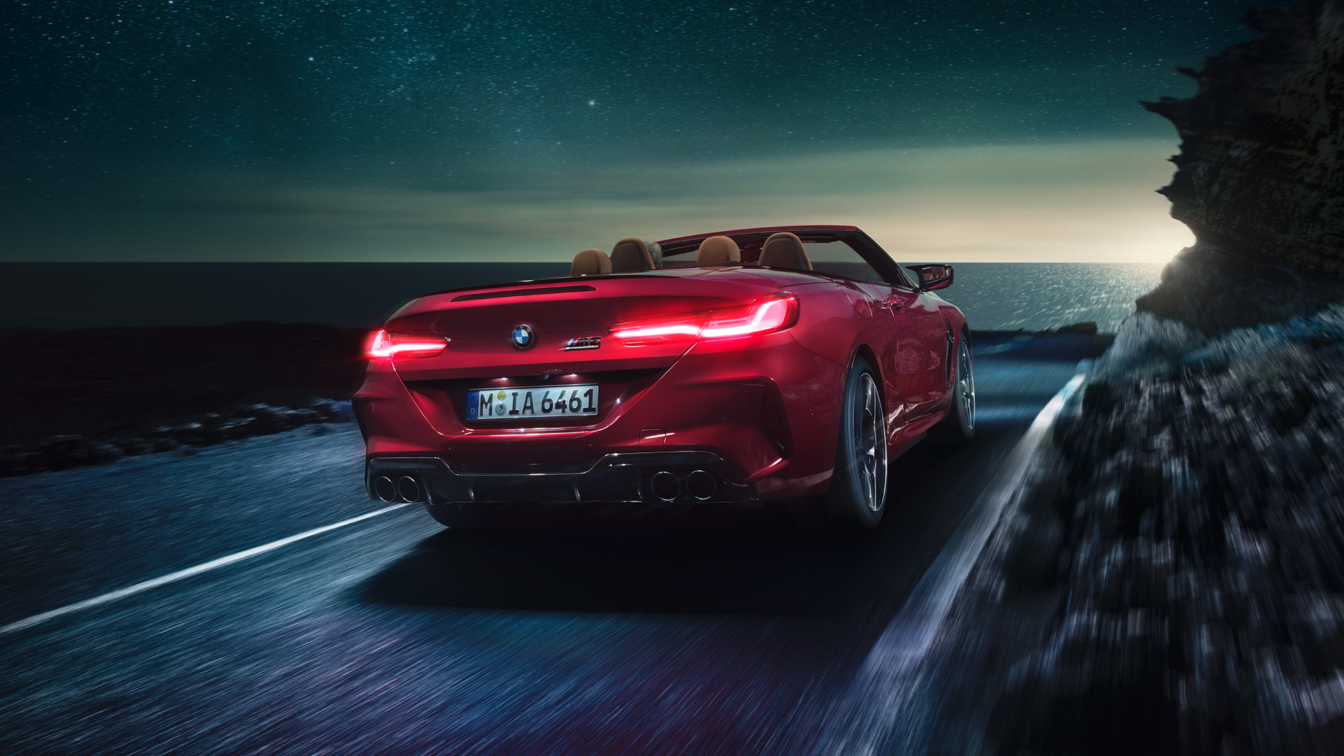 BMW M8 Competition Cabriolet set bagfra let drejet. BMW M8 Competition Cabriolet i Motegi Red metallic med åben kaleche set bagfra let drejet, kørende om natten på en kystvej.