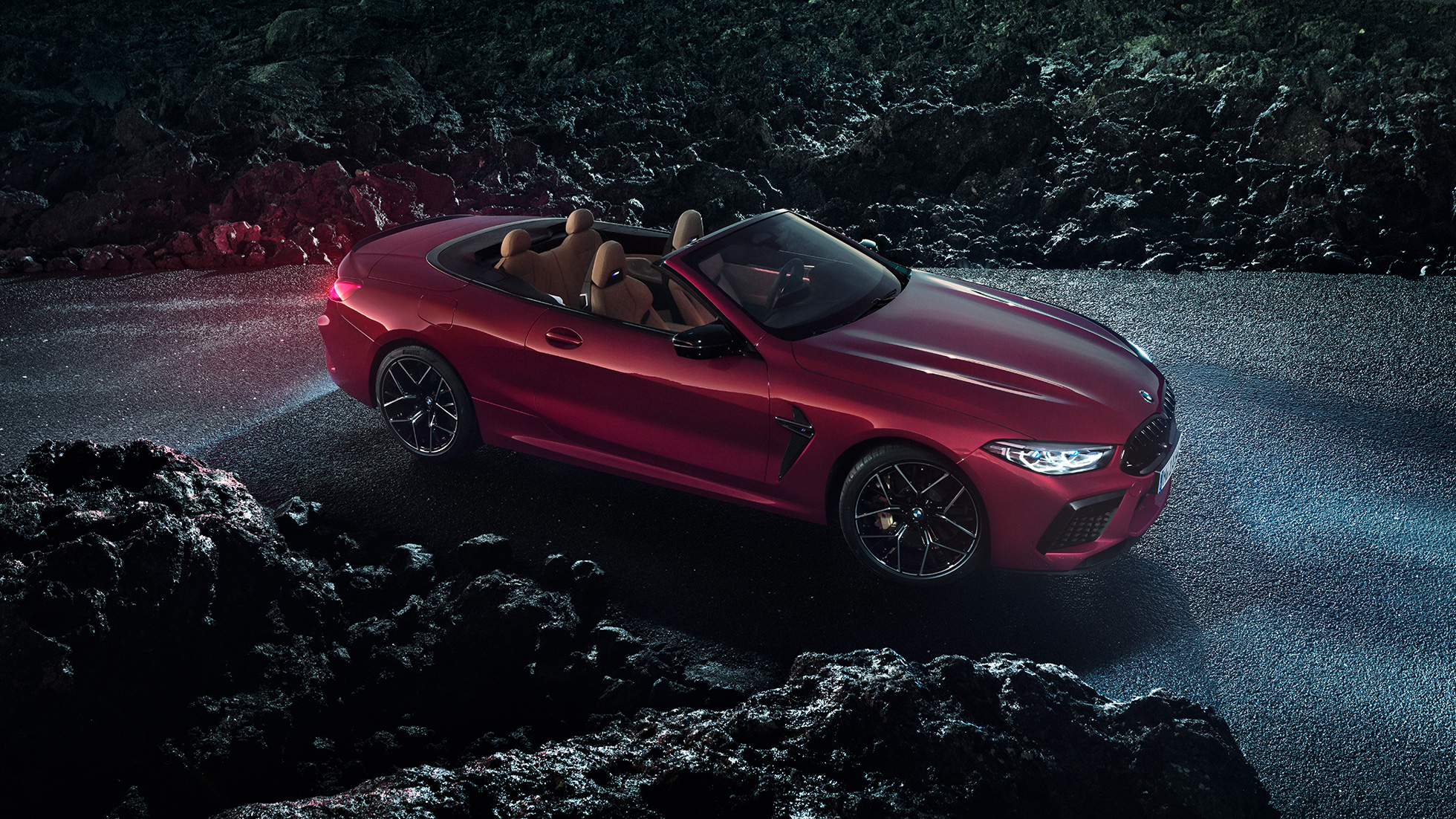 BMW M8 Competition Cabriolet set fra siden let drejet. BMW M8 Competition Cabriolet i Motegi Red metallic med åben kaleche set fra siden let drejet, parkeret om natten på asfalt.