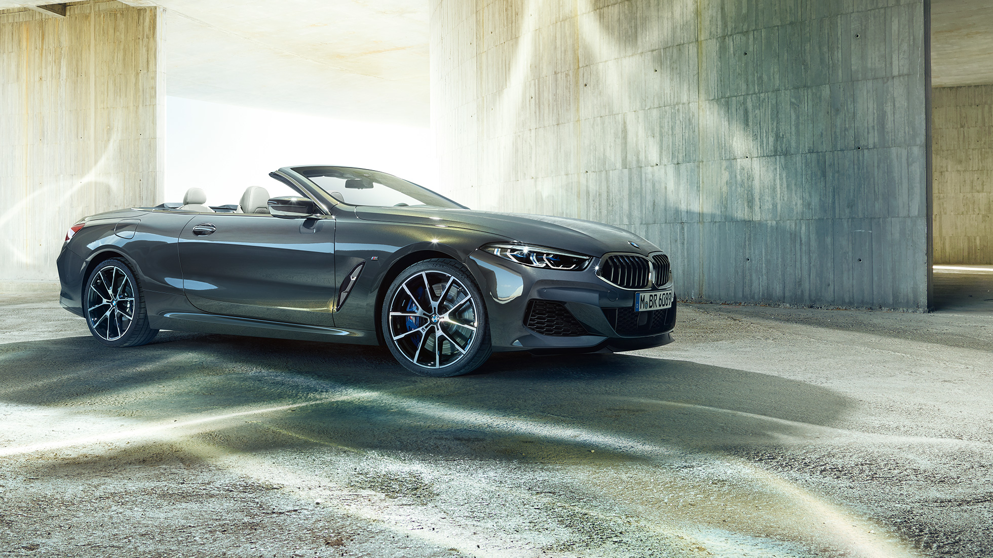 BMW M850i xDrive Cabriolet set fra siden let drejet. BMW M850i xDrive Cabriolet i Dravit Grey metallic med åben kaleche set fra siden let drejet, parkeret foran en betonbygning.