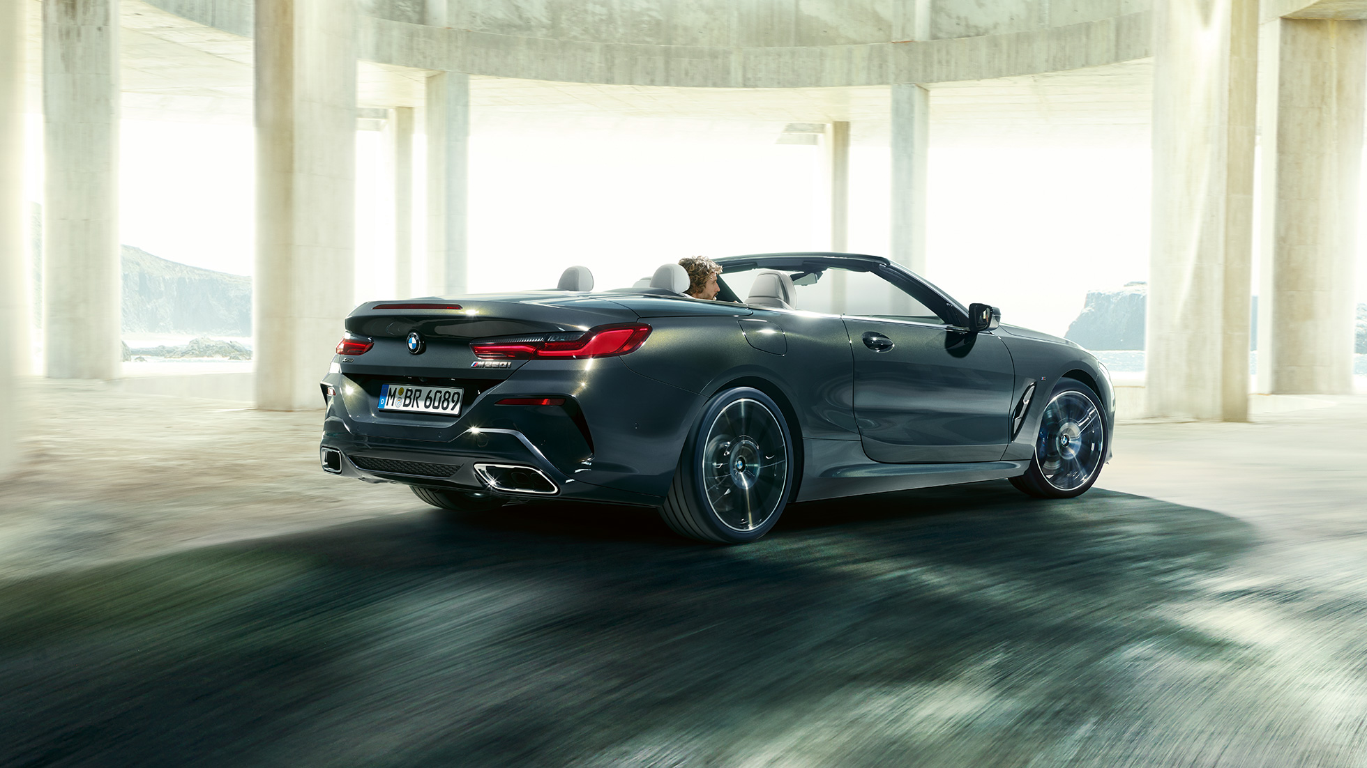BMW M850i xDrive cabriolet set bagfra let drejet. BMW M850i xDrive Cabriolet i Dravit Grey metallic med åben kaleche set bagfra let drejet kørende gennem en betonbygning.