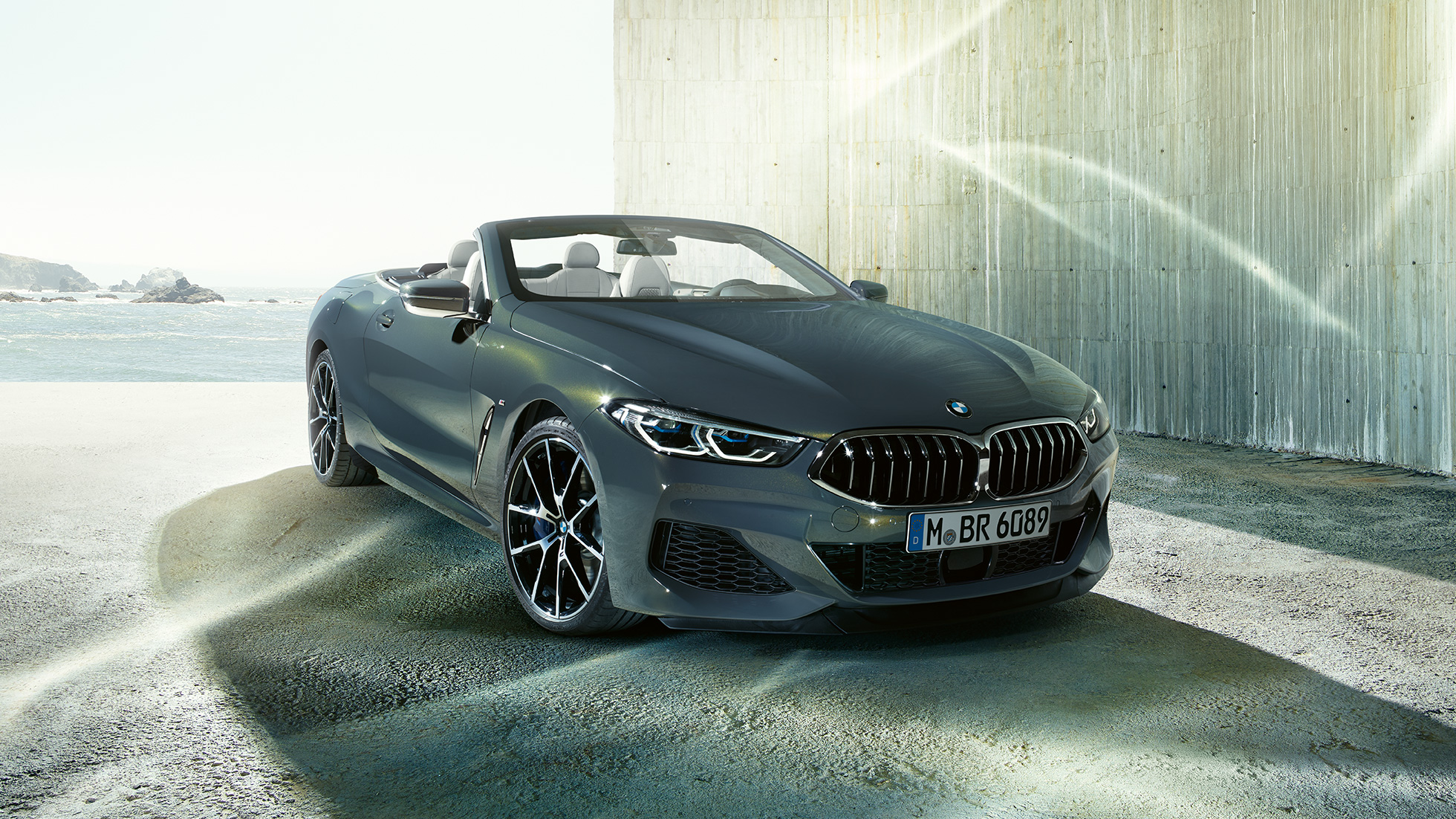 BMW M850i xDrive Cabriolet set forfra let drejet. BMW M850i xDrive Cabriolet i Dravit Grey metallic med åben kaleche set forfra let drejet, parkeret foran en kyst.