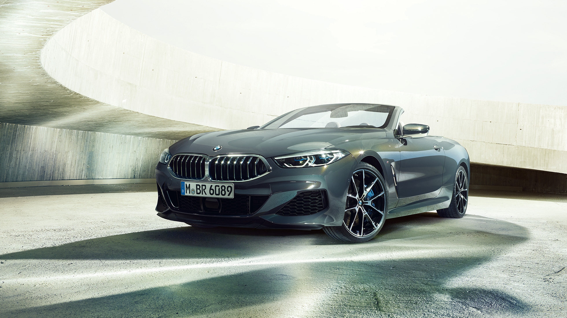 BMW M850i xDrive Cabriolet set forfra let drejet. BMW M850i xDrive Cabriolet i Dravit Grey metallic med åben kaleche set forfra let drejet, parkeret i en betonbygning.