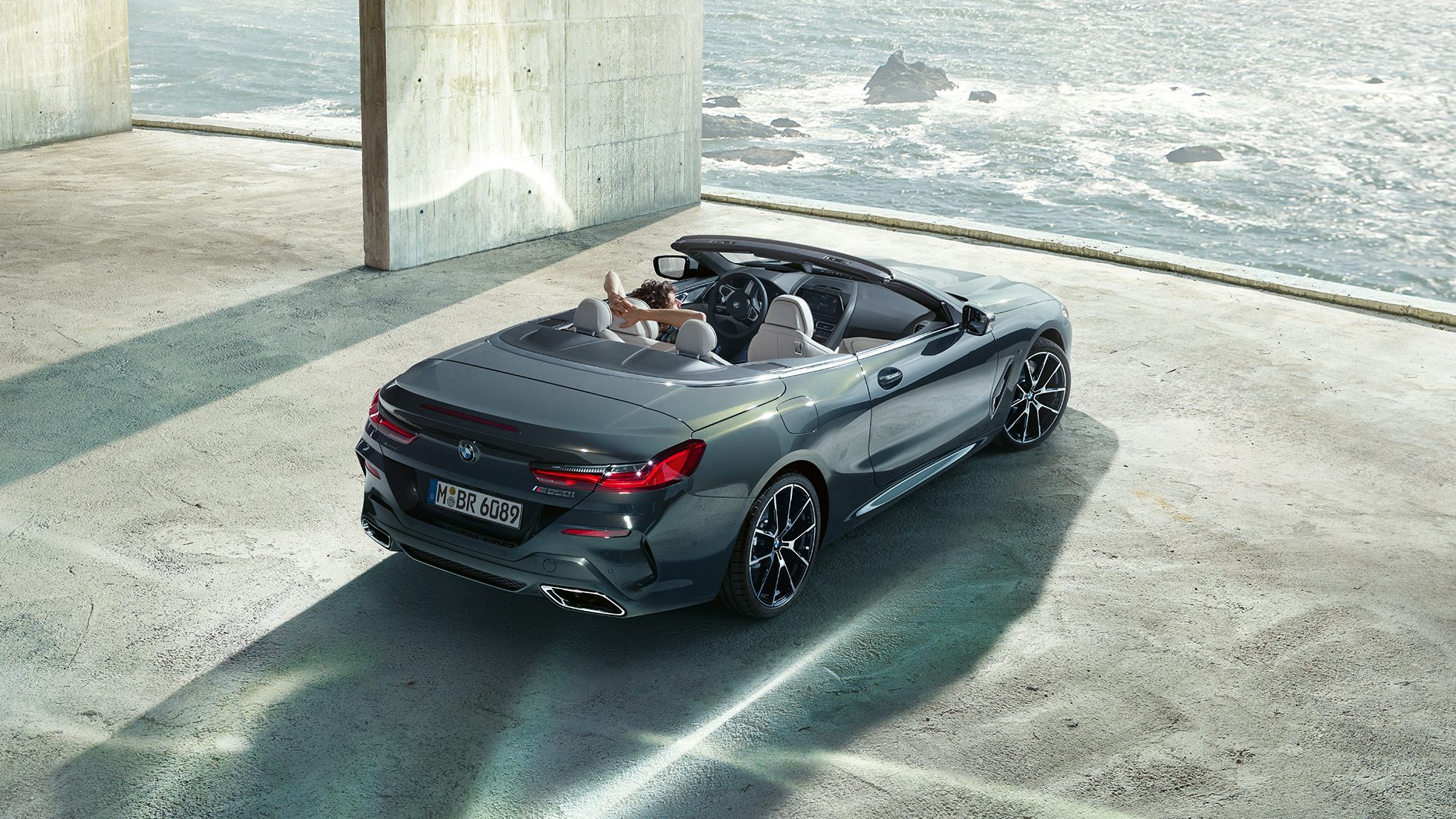 BMW M850i xDrive Cabriolet i fugleperspektiv. BMW M850i xDrive Cabriolet i Dravit Grey metallic med åben kaleche set i fugleperspektiv, mens den parkerer i en betonbygning foran et ocean.