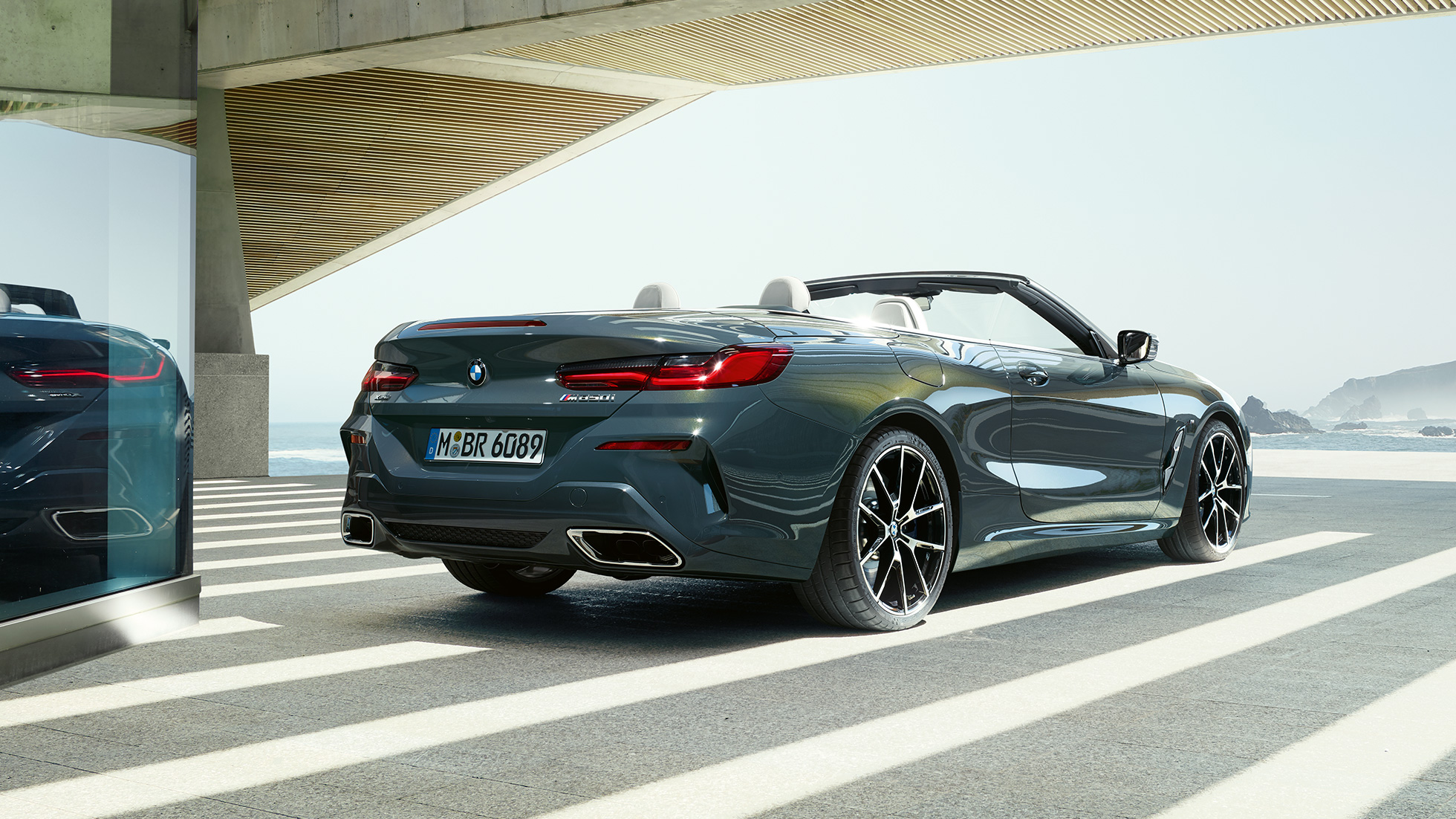 BMW M850i xDrive cabriolet set bagfra let drejet. BMW M850i xDrive Cabriolet i Dravit Grey metallic med åben kaleche set bagfra let drejet, mens den parkerer ud mod et ocean.