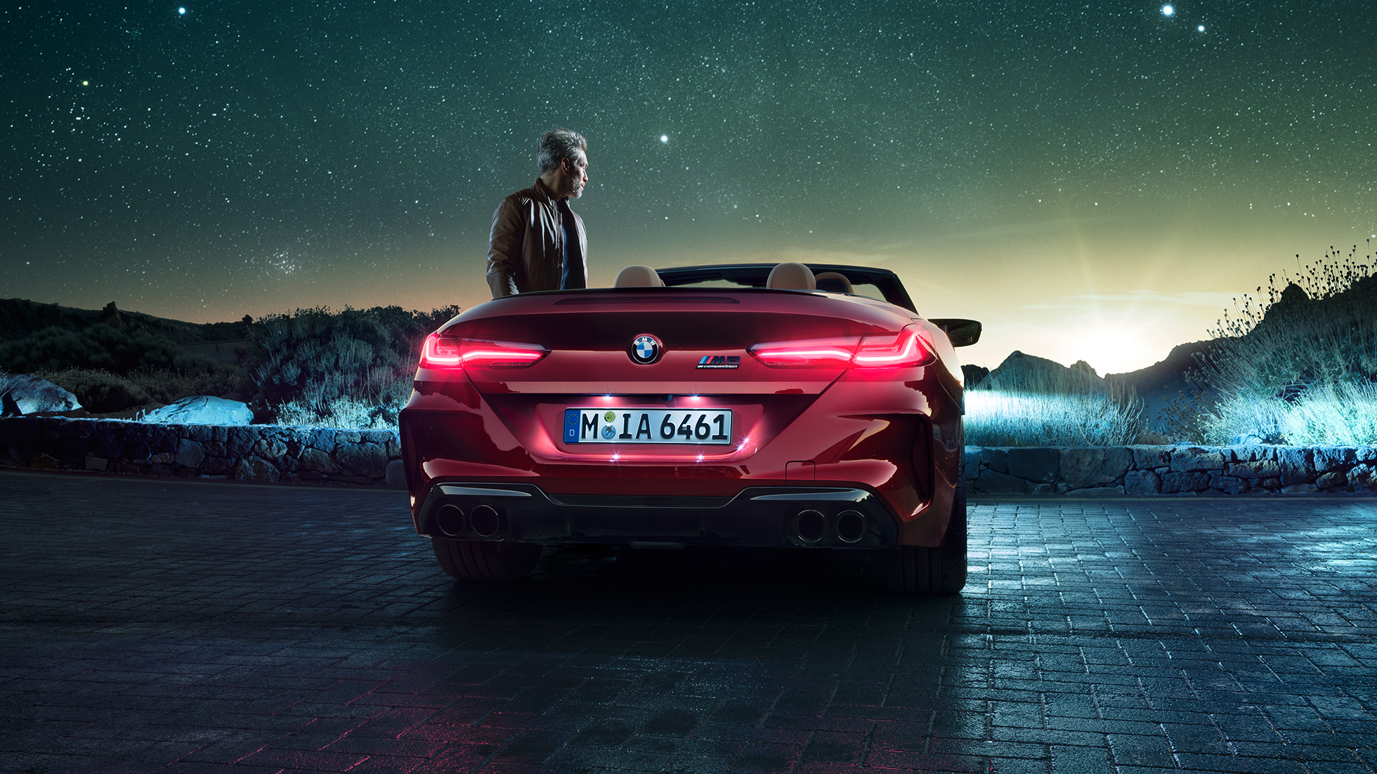 BMW M8 Competition Cabriolet set bagfra. BMW M8 Competition Cabriolet i Motegi Red metallic med åben kaleche set bagfra ved siden af en mand under en stjernehimmel.