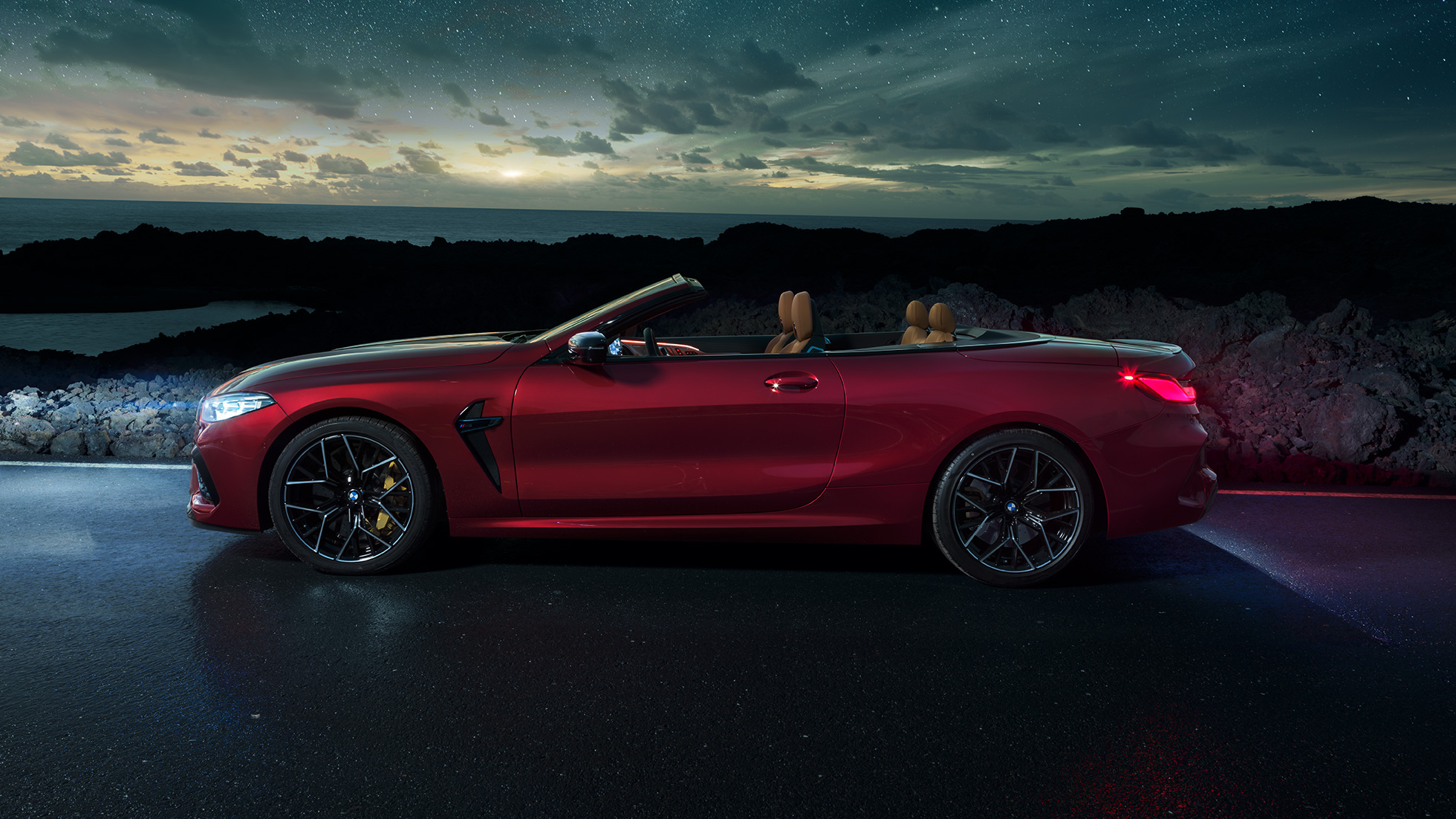 BMW M8 Competition Cabriolet set fra siden. BMW M8 Competition Cabriolet i Motegi Red metallic med åben kaleche set fra siden parkeret om natten under en stjernehimmel.