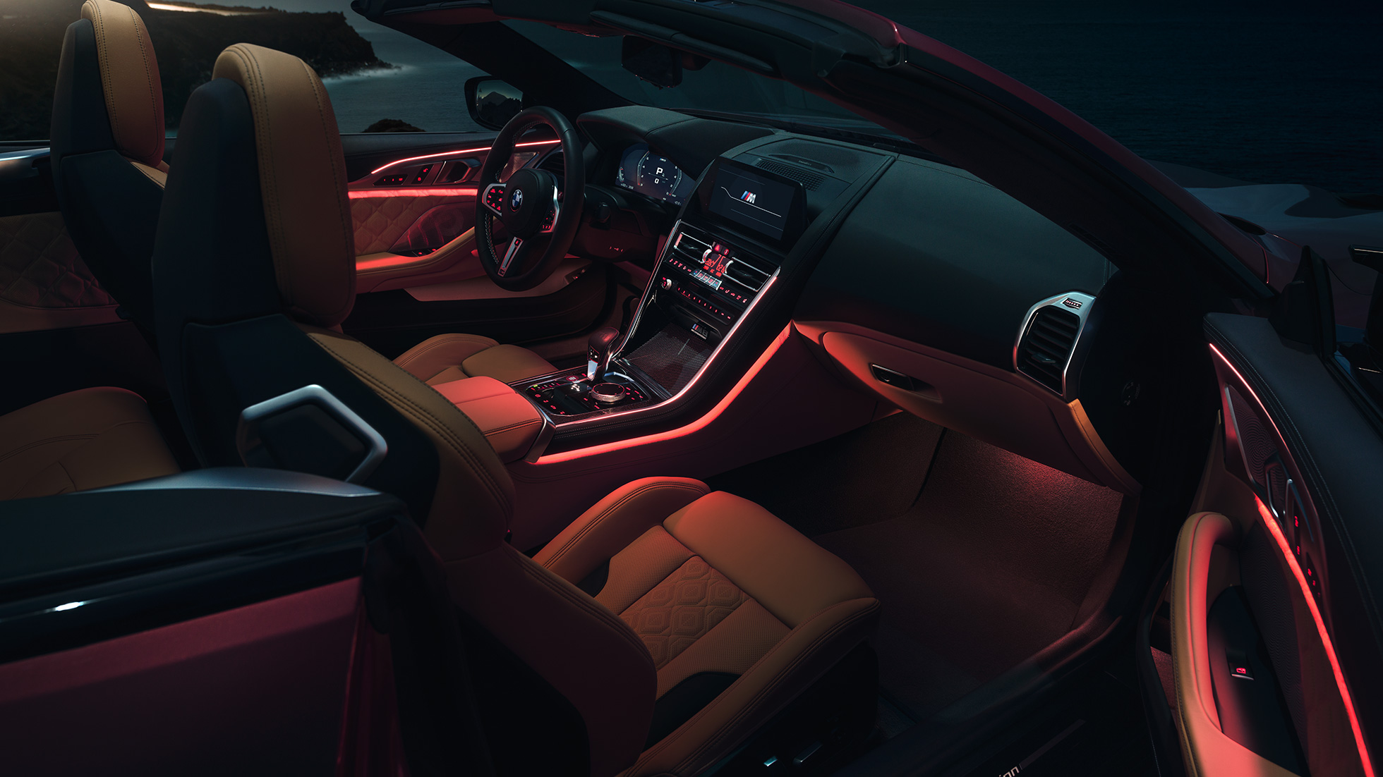 BMW M8 Competition Cabriolet, interiør. BMW M8 Competition Cabriolet med åben kaleche og rødt Ambient Light-kabinelys om natten.