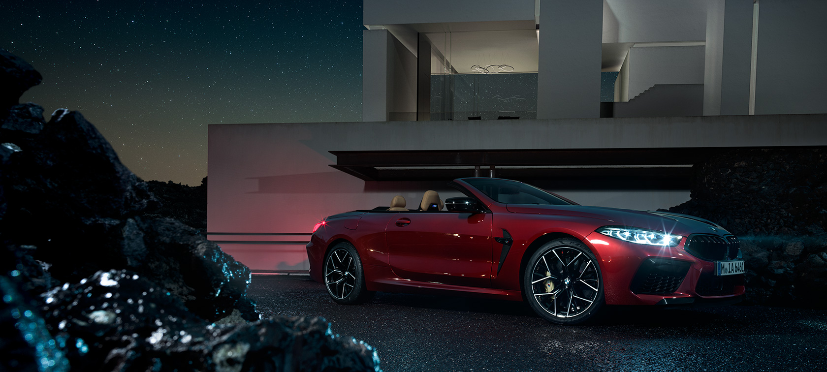 BMW M8 Competition Cabriolet set fra siden let drejet. BMW M8 Competition Cabriolet i Motegi Red metallic, set fra siden let drejet, parkeret om natten foran en villa.