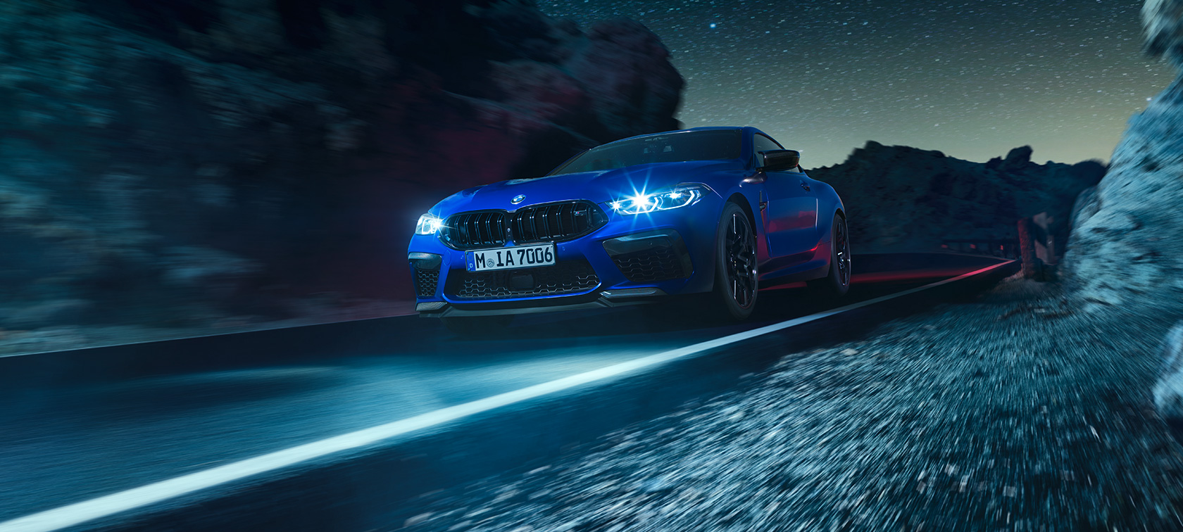 BMW M8 Competition Coupé set forfra let drejet. BMW M8 Competition Coupé i Marina Bay Blue metallic set forfra let drejet, kørende om natten.