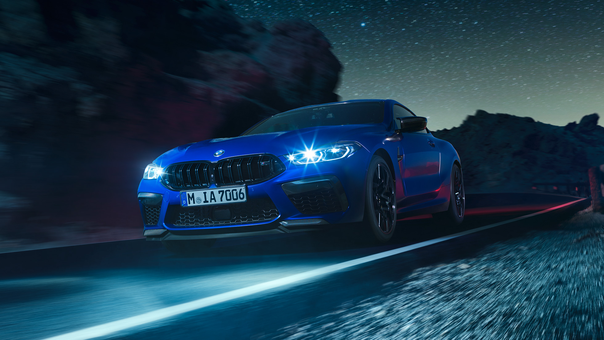 Kørende BMW M8 Competition Coupé set forfra let drejet. BMW M8 Competition Coupé i Marina Bay Blue metallic med skinnende LED-forlygter set forfra let drejet, kørende om natten.