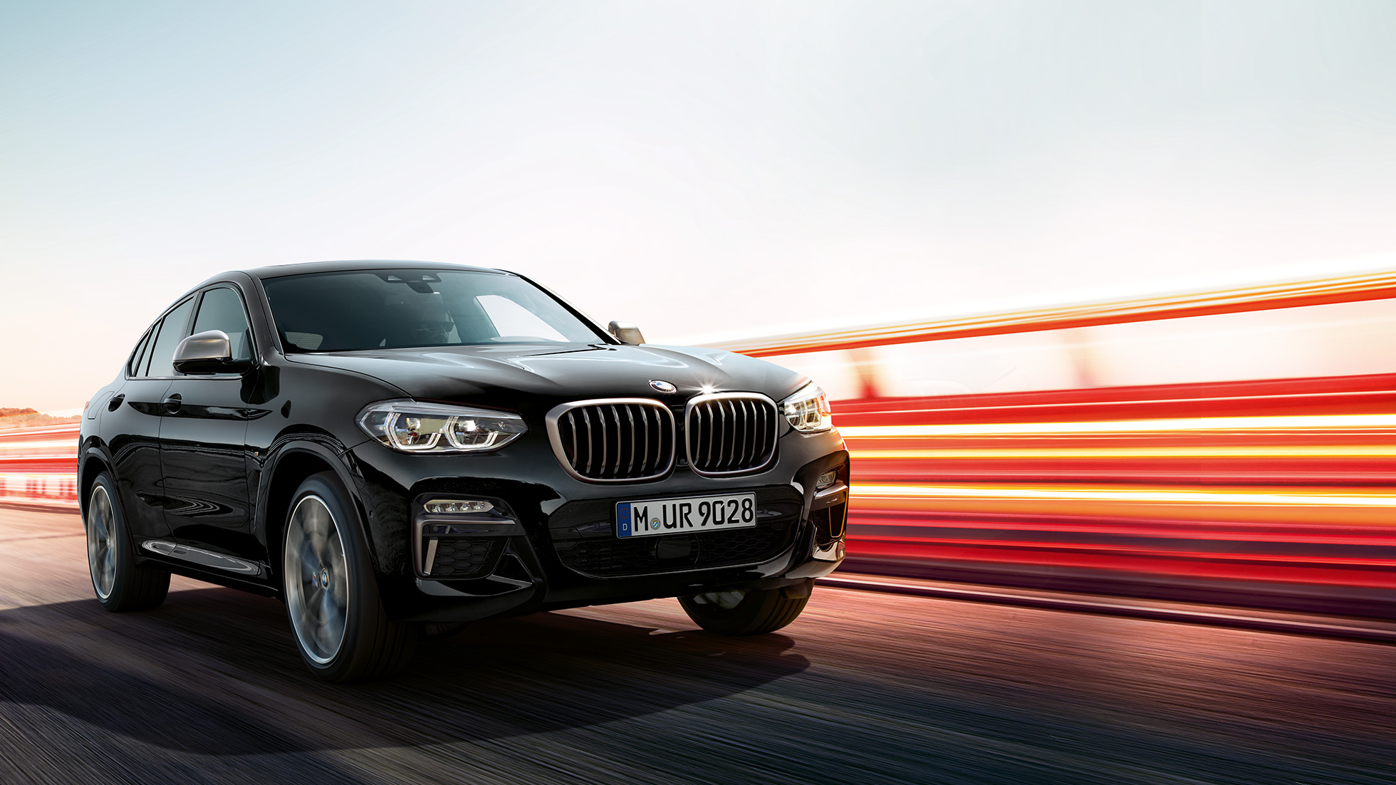 BMW X4 M40i, set bagfra let drejet. BMW X4 M40i i safirsort metallic, kørende, eksteriør, set bagfra let drejet med lysstriber.