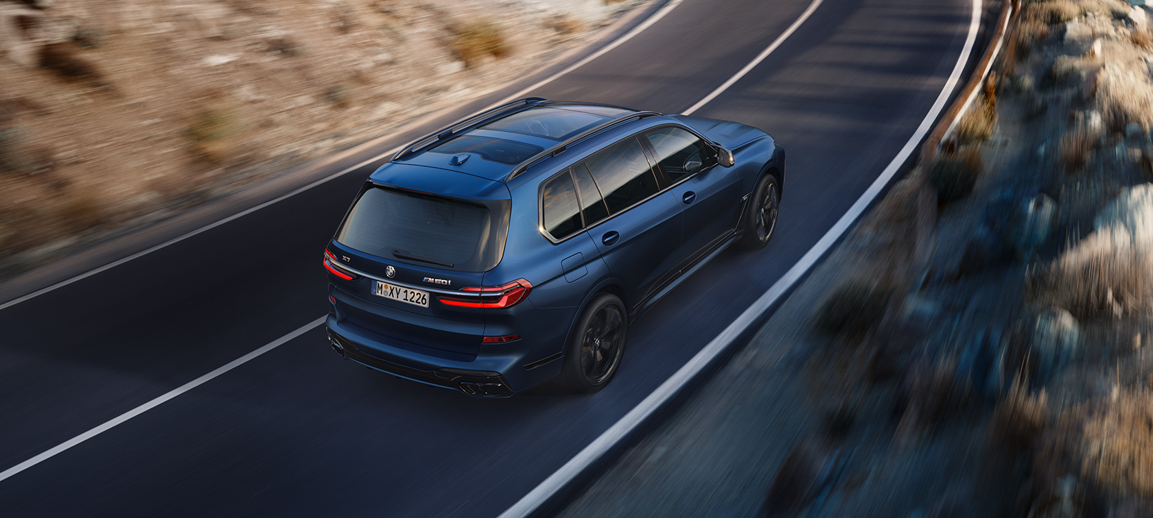 BMW X7 M60i xDrive G07 LCI Frozen Marina Bay Blue set bagfra let drejet i fugleperspektiv kørende BMW X7 M60i xDrive G07 LCI Frozen Marina Bay Blue set bagfra let drejet i fugleperspektiv kørende på en vej
