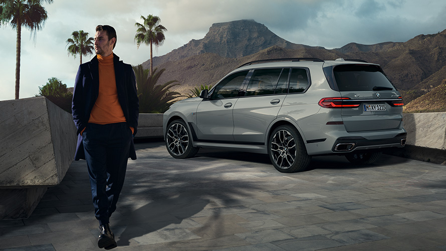 En mand foran en BMW X7 (G07) Remote Control Parking Remote Control Parking en mand foran en parkeret BMW X7 (G07) set bagfra let drejet under palmer med et bjerglandskab i baggrunden
