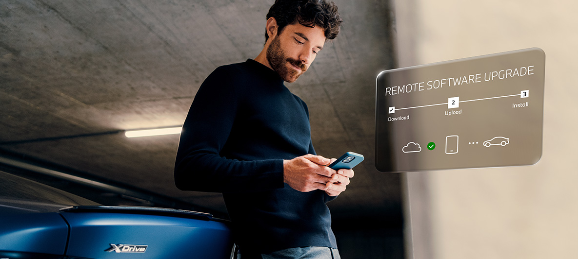 En mand læner sig op ad hækken på en BMW og kigger på sin smartphone Remote Software Upgrade en mand læner sig op ad hækken på en BMW og kigger på sin smartphone