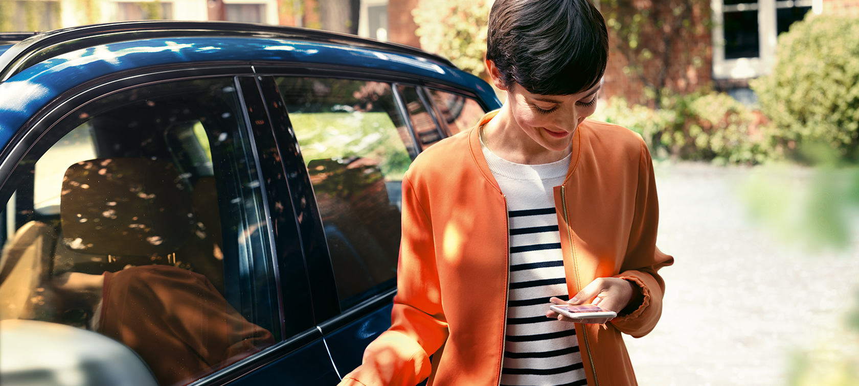 Mørkhåret kvinde foran BMW Mørkhåret kvinde i orange jakke står foran en BMW og kigger på sin smartphone