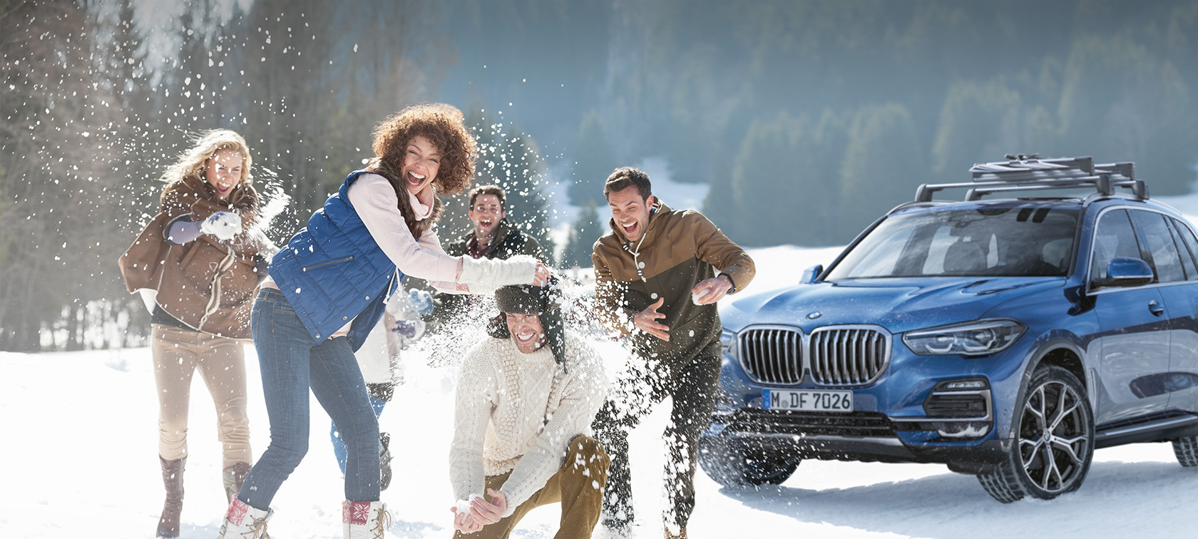 Originalt BMW tilbehør og BMW service til efterår og vinter En gruppe kvinder og mænd griner i sneen foran en BMW