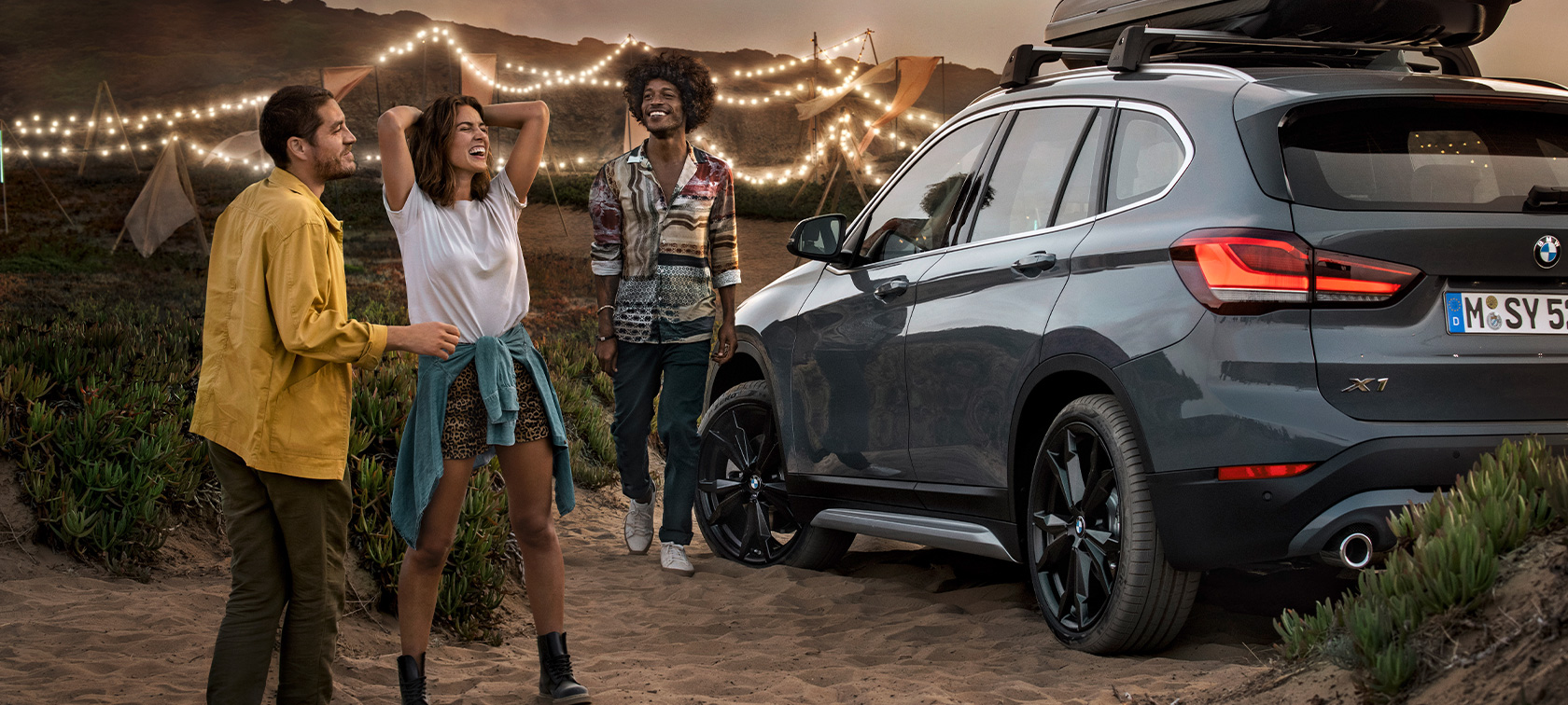En jublende trio i en BMW X1 med BMW sommertilbud En lykkelig trio i en Mineral Grey BMW X1 udstyret med BMW sommertilbud set bagfra let drejet på en strand