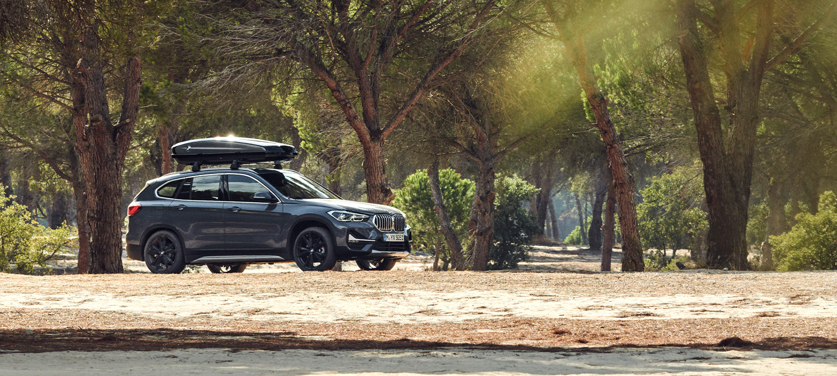 BMW X1 set fra siden med tagboks BMW sommertilbud BMW X1 i Mineral Grey set fra siden med BMW tagboks parkeret offroad i en skov BMW sommertilbud