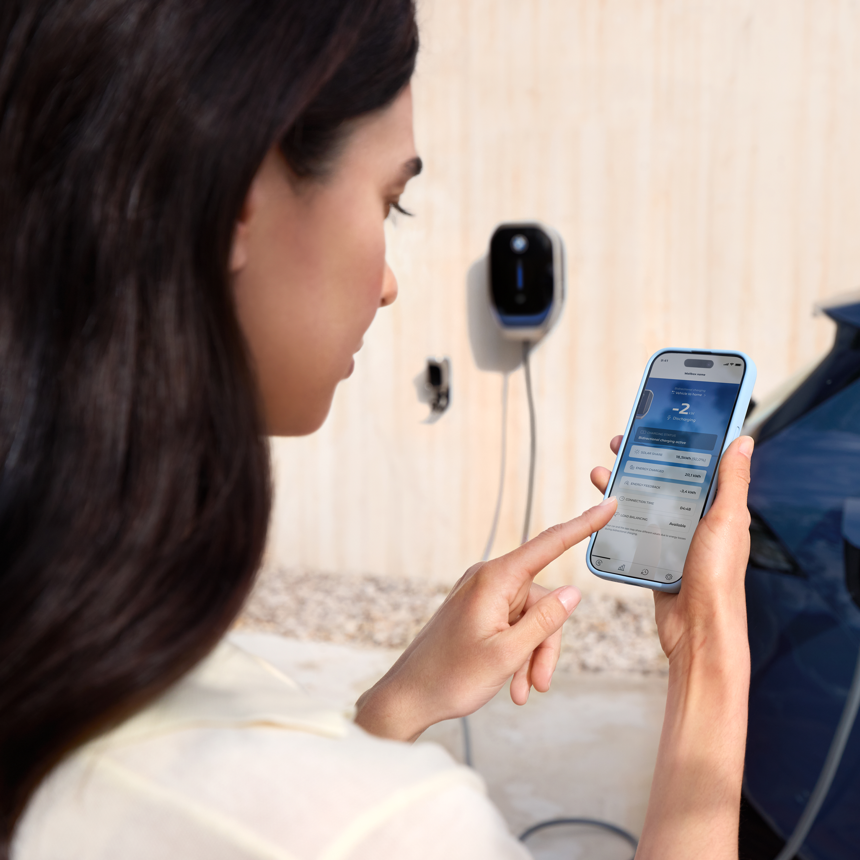En kvinde ser på sin smartphone, en wallbox er synlig i baggrunden